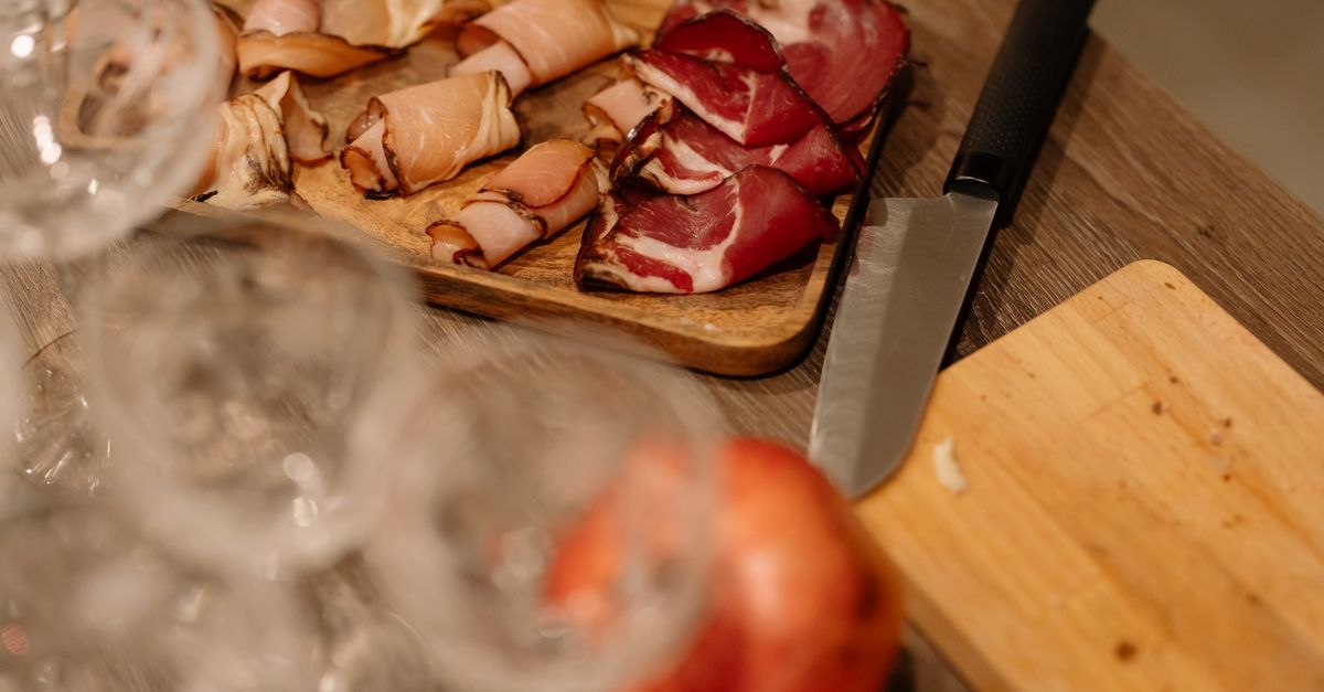 Tabla de madera con lonchas de jamón curado y otros embutidos, junto a un cuchillo y copas de vino en primer plano.