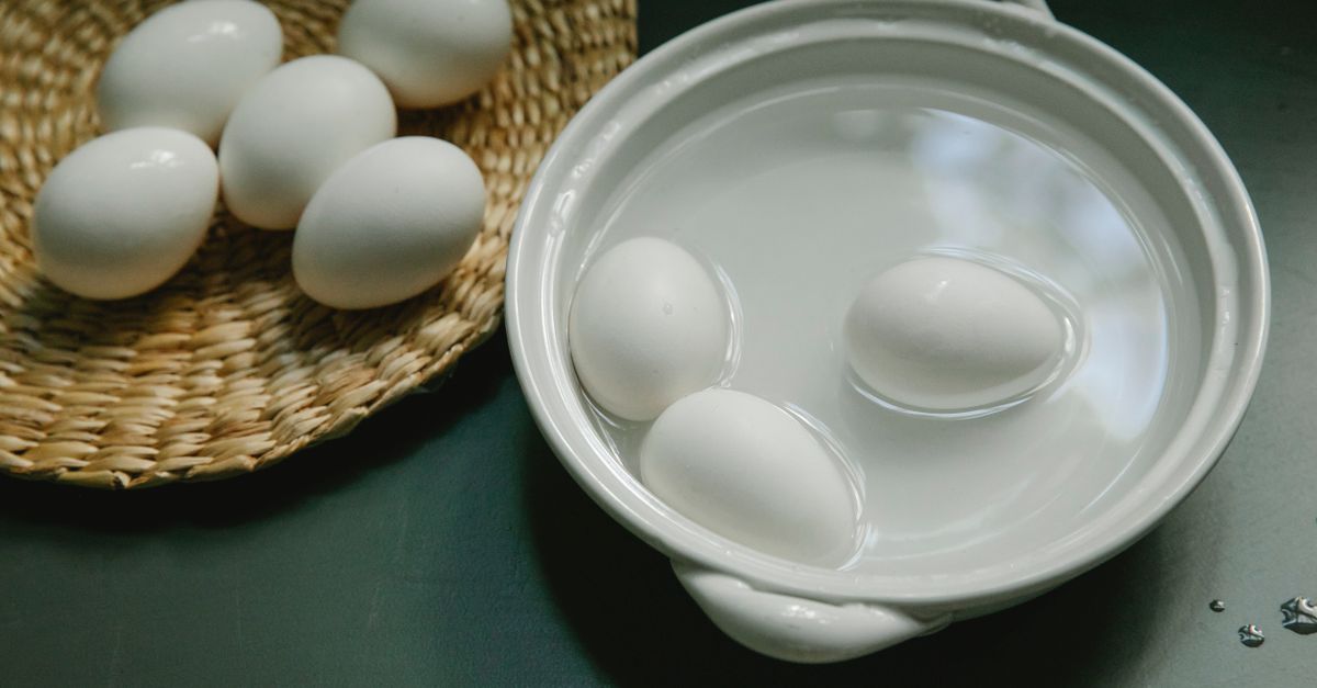 Tres huevos blancos sumergidos en agua dentro de un recipiente de cerámica, junto a otros huevos en un cesto de mimbre.