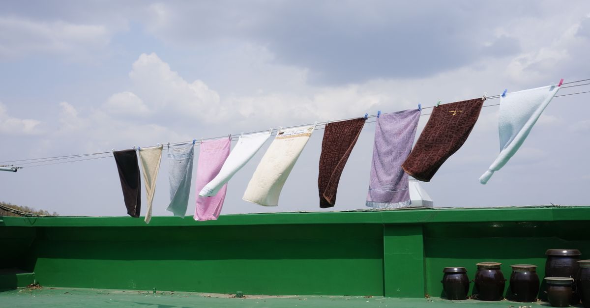 Varias toallas y prendas de colores cuelgan de una cuerda de tender al aire libre bajo un cielo nublado, con tinajas de barro al fondo.
