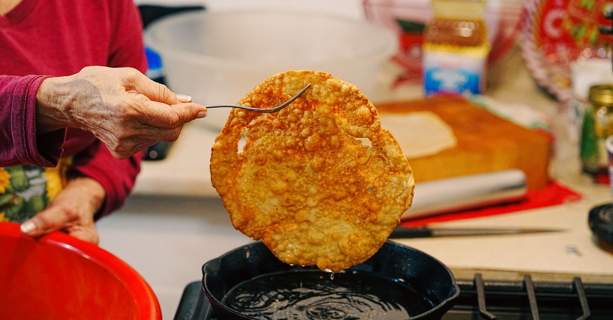 Una persona levanta con un tenedor una tortilla o filete dorado y crujiente desde una sartén con aceite sobre el fuego.
