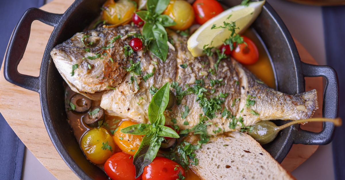 Un pescado entero cocinado en una sartén de hierro fundido, acompañado de tomates cherry, aceitunas, hierbas frescas y una rodaja de limón, con un trozo de pan al lado.