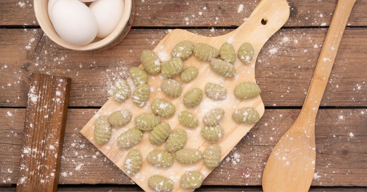 Ñoquis o gnocchi caseros de color verde dispuestos sobre una tabla de madera con forma de pala, junto a un cuenco con huevos, un rodillo y una cuchara de madera con harina espolvoreada.