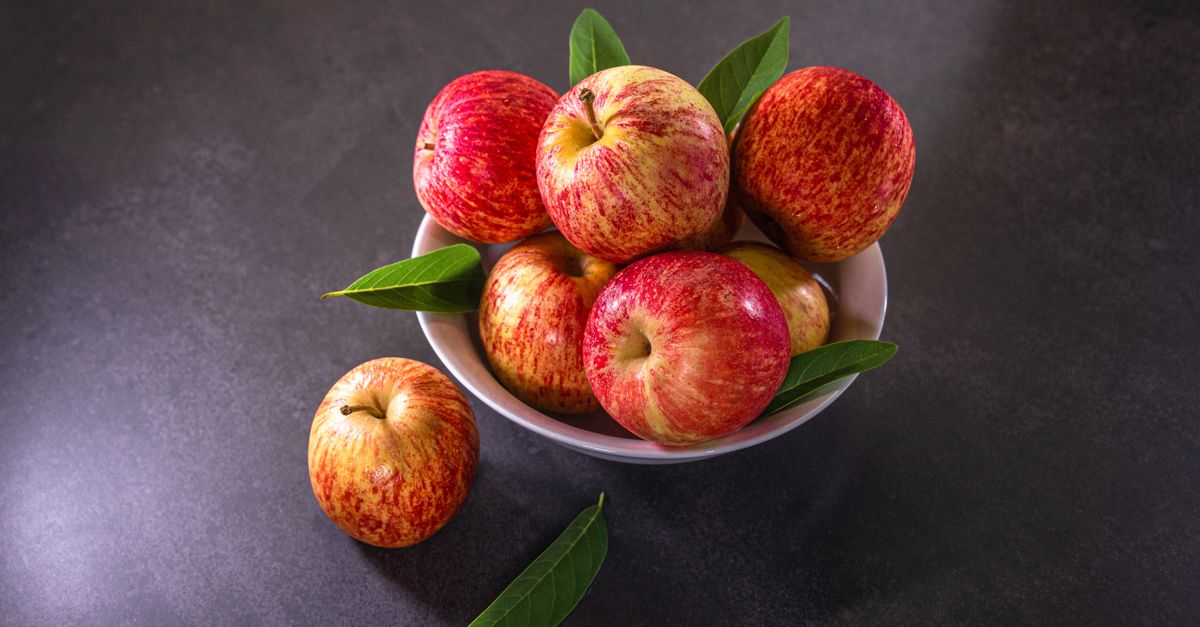 Un bol blanco lleno de manzanas rojas y amarillas frescas con hojas verdes, sobre una superficie oscura, con una manzana adicional al lado.