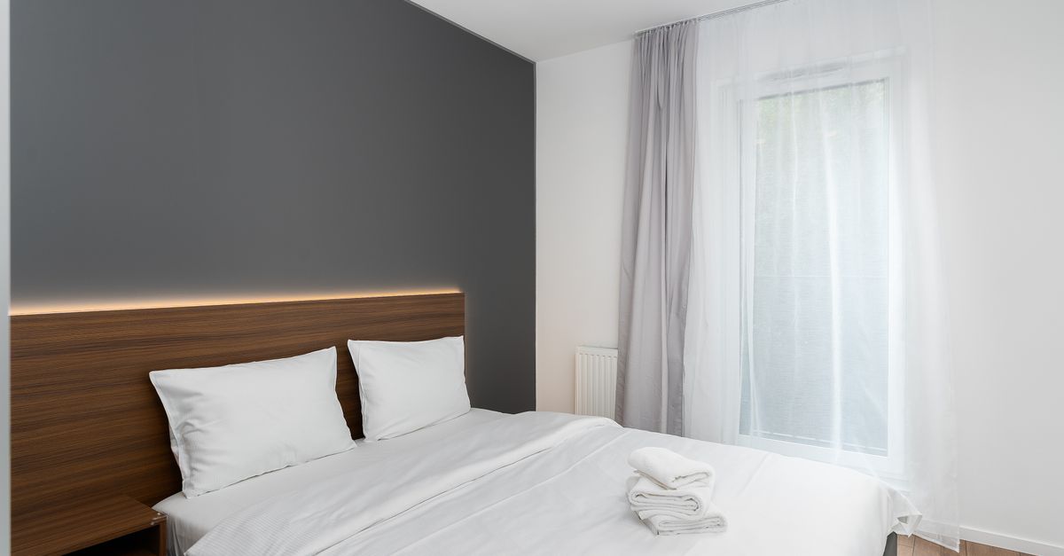 Dormitorio ordenado con cama de sábanas blancas bien hechas, cabecero de madera y ventana con cortina translúcida.