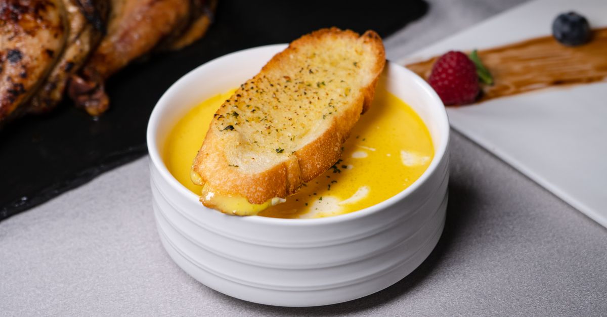 Un bol blanco de crema de verduras color amarillo con una tostada de pan crujiente apoyada encima, acompañado de fruta y otros elementos decorativos alrededor.