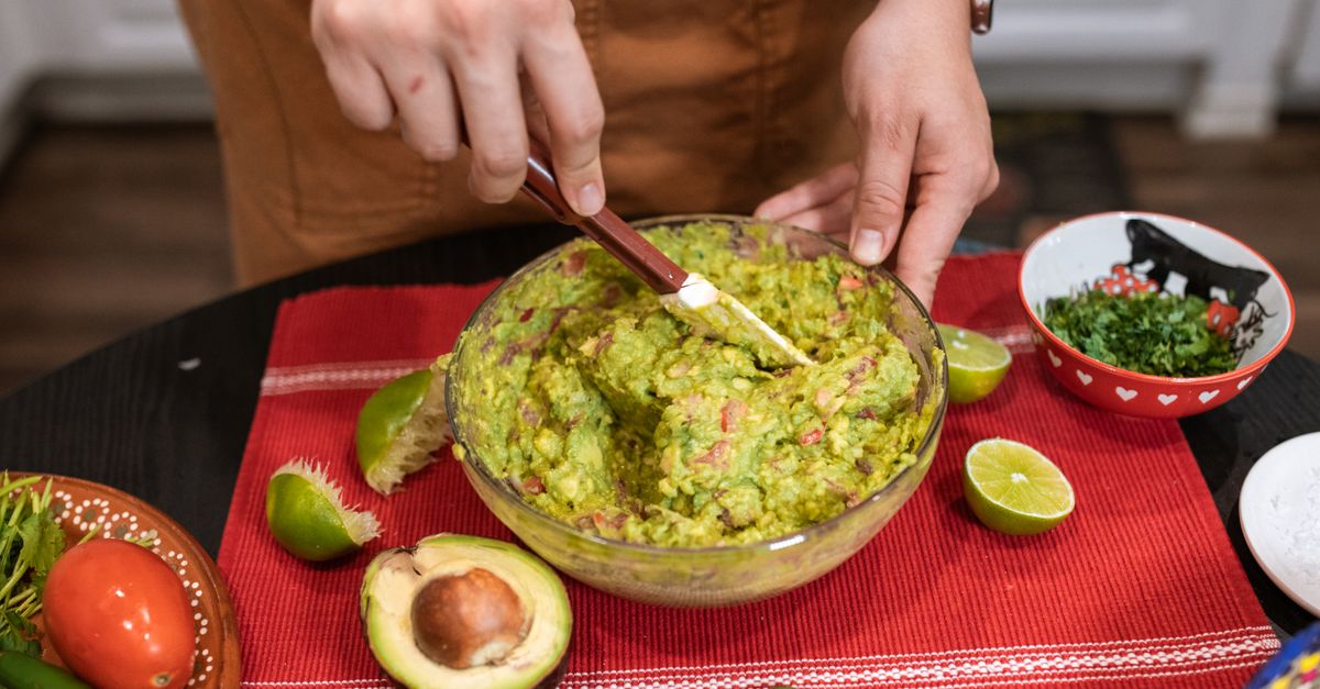 Un cuenco de guacamole recién preparado sobre un mantel rojo, rodeado de limas cortadas y un aguacate partido, mientras una persona lo mezcla con una cuchara.