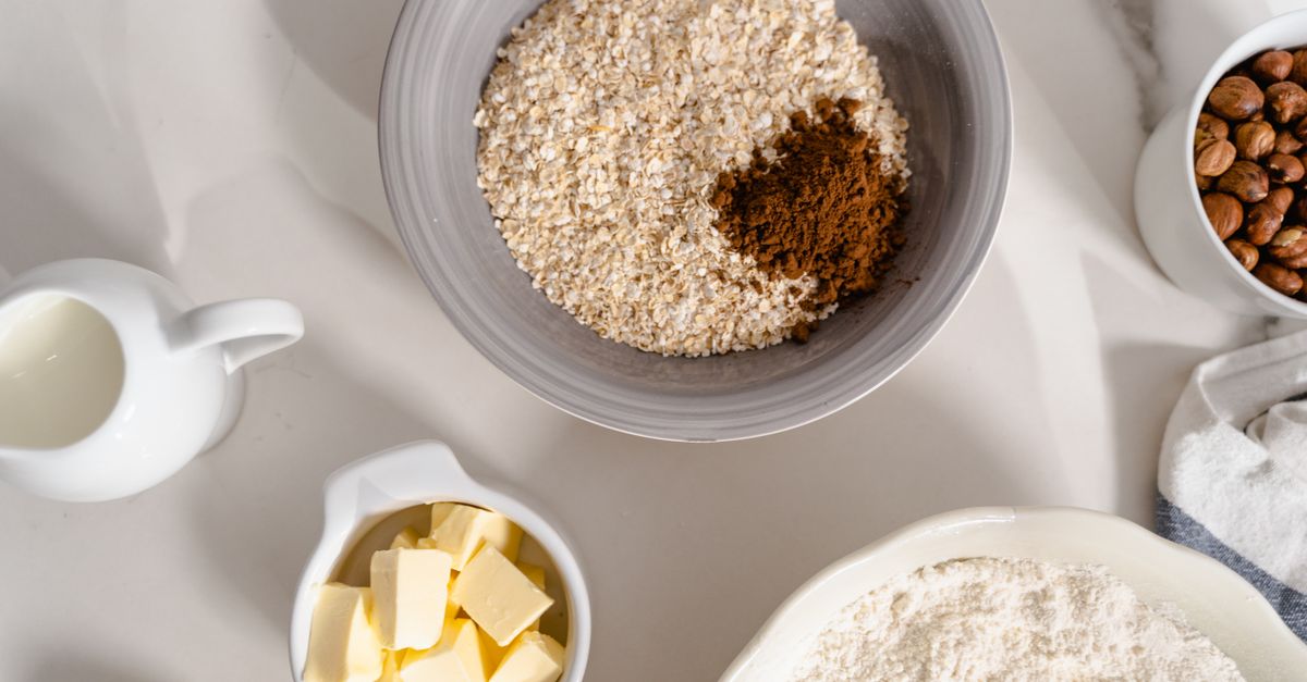 Un bowl con avena y canela junto a un pequeño recipiente con cubos de mantequilla, harina y otros ingredientes sobre una encimera blanca.