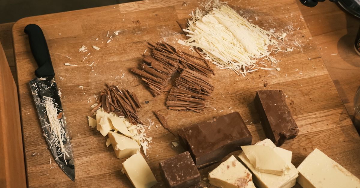 Tabla de madera con virutas de queso rallado, tiras de chocolate y trozos de mantequilla y chocolate blanco cortados en cubos, junto a un cuchillo.