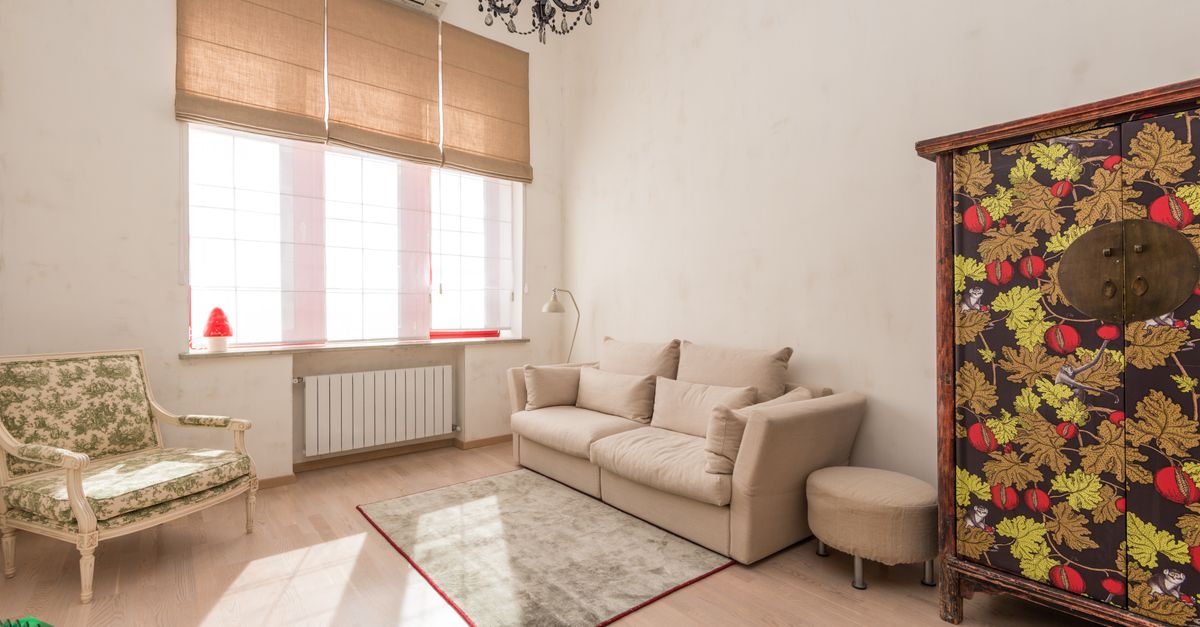 Salón acogedor con radiador blanco bajo una ventana con estor, sofá beige y sillón vintage junto a una alfombra.