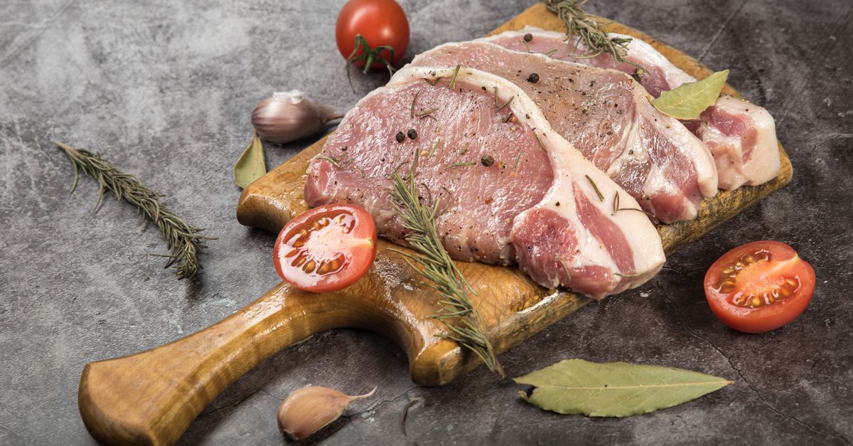 Cortes de carne cruda de cerdo dispuestos sobre una tabla de madera junto a tomates, ajos, romero y hojas de laurel.