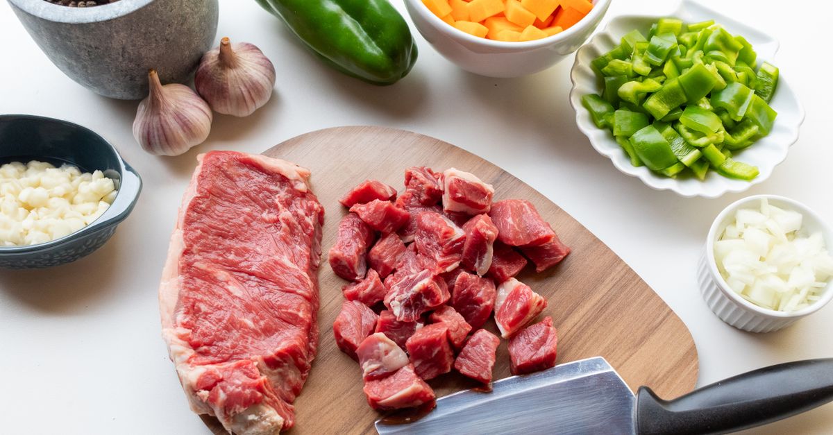 Un trozo de carne cruda entera junto a carne cortada en cubos sobre una tabla de madera, rodeada de cuencos con ajo, pimiento verde, zanahoria y cebolla picados y un cuchillo cebollero.