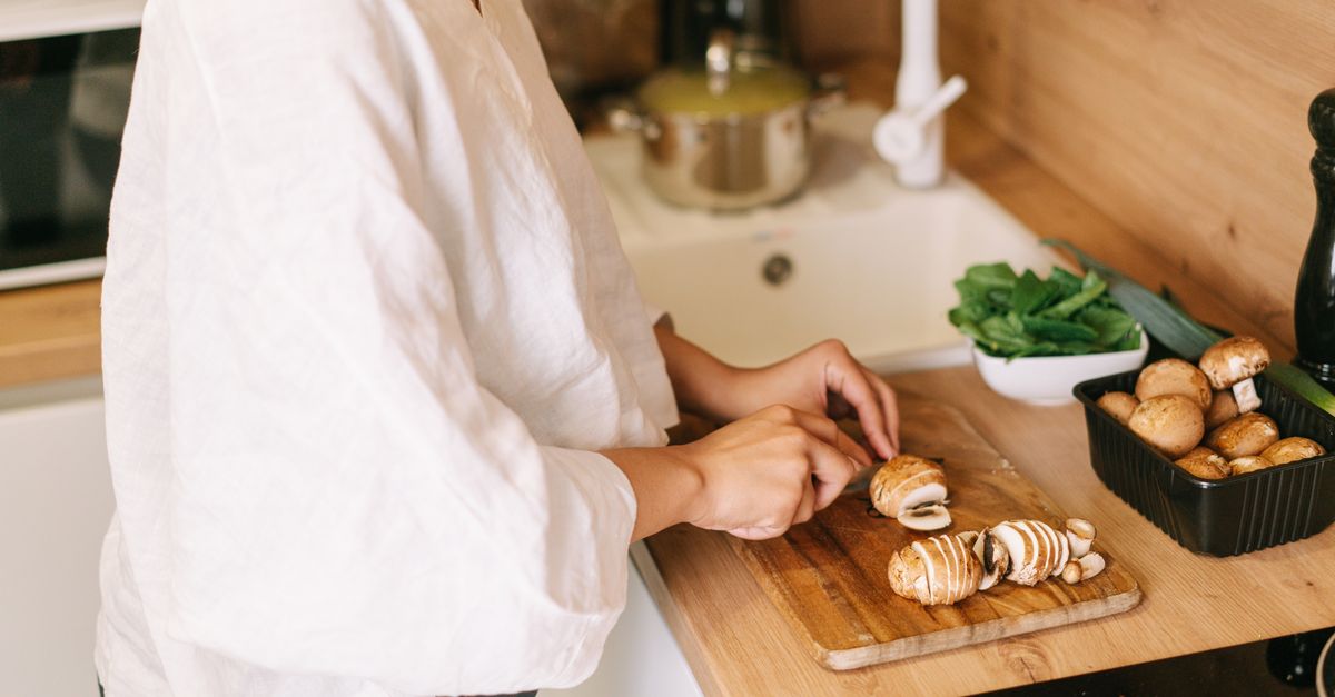 Una persona corta champiñones frescos sobre una tabla de madera en una encimera de cocina, junto a un envase con más champiñones y verduras.