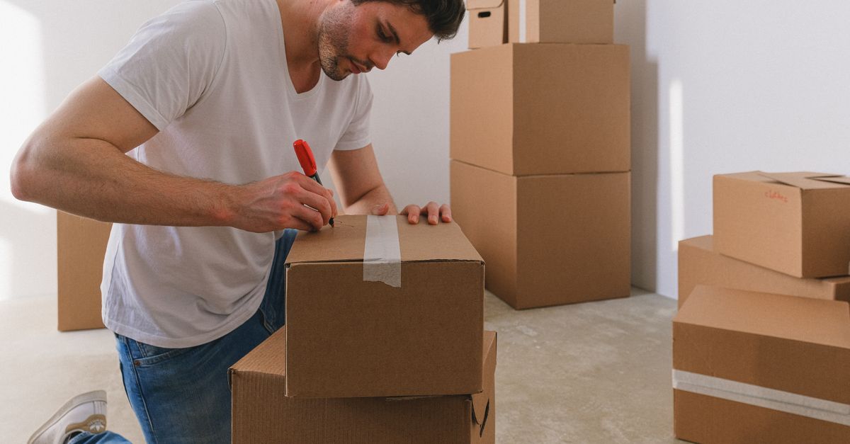 Un hombre escribe sobre una caja de cartón con un marcador rojo, rodeado de varias cajas apiladas en una habitación.