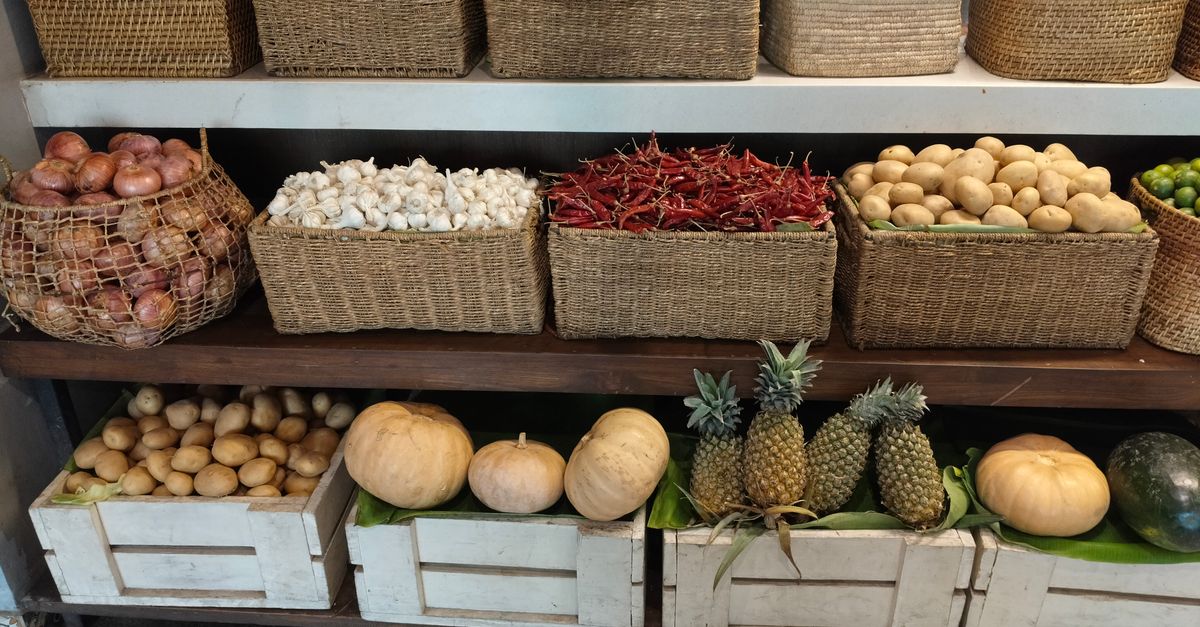 Cestas de mimbre con patatas y otras verduras dispuestas en estanterías de madera en un rincón de despensa con poca luz.