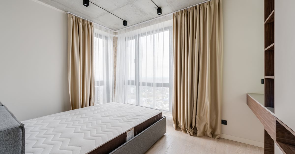 Colchón desnudo sobre una base de madera en un dormitorio con grandes ventanales y cortinas abiertas dejando entrar luz natural.