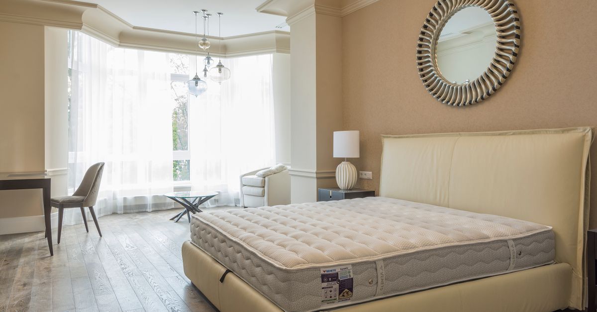 Colchón desnudo sobre una base de cama en un dormitorio luminoso con ventanales y cortinas blancas.