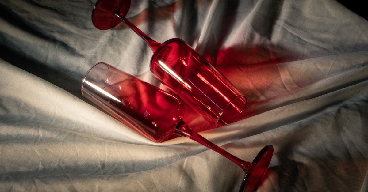 Dos copas rojas volcadas sobre una sábana blanca arrugada, con la luz proyectando un reflejo rojizo intenso sobre la tela.