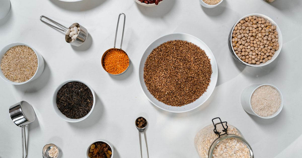 Vista cenital de varios cuencos con legumbres, arroz y cereales junto a tazas y cucharas medidoras metálicas sobre una superficie blanca.