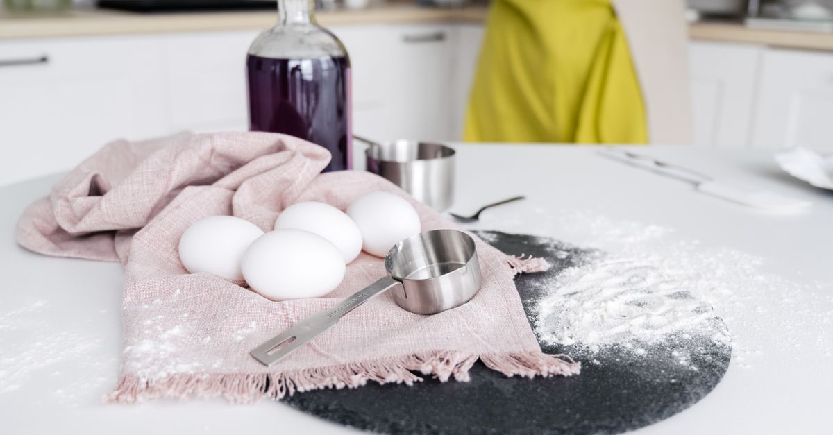 Mesa de cocina con huevos, una botella morada, un paño rosa y una taza medidora metálica junto a harina esparcida sobre la encimera.