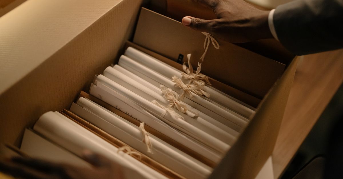 Una mano organiza carpetas de papel atadas con cuerdas dentro de una caja de cartón sobre un escritorio de madera.