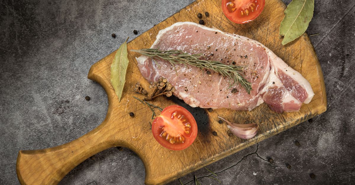Un filete de carne cruda sobre una tabla de madera, acompañado de tomate cortado, romero, ajo y pimienta.