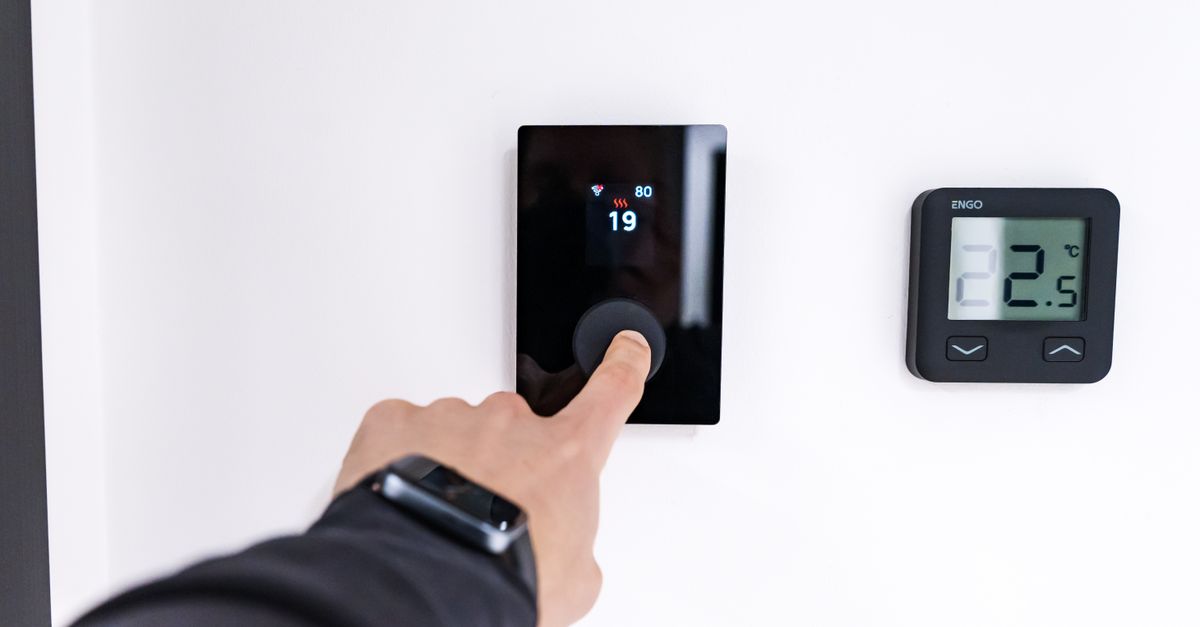 Una mano ajusta un termostato digital de pared marcando 19 grados mientras otro termostato con pantalla muestra 22.5 grados al lado.