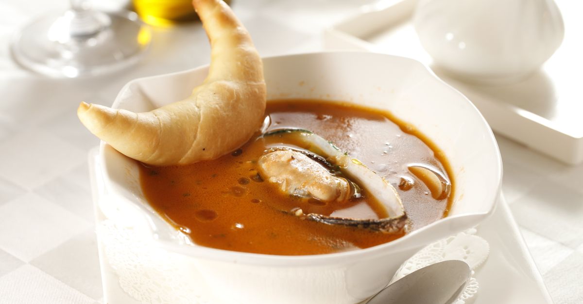 Un cuenco blanco de sopa de pescado con mejillones y un trozo de pan crujiente apoyado en el borde.