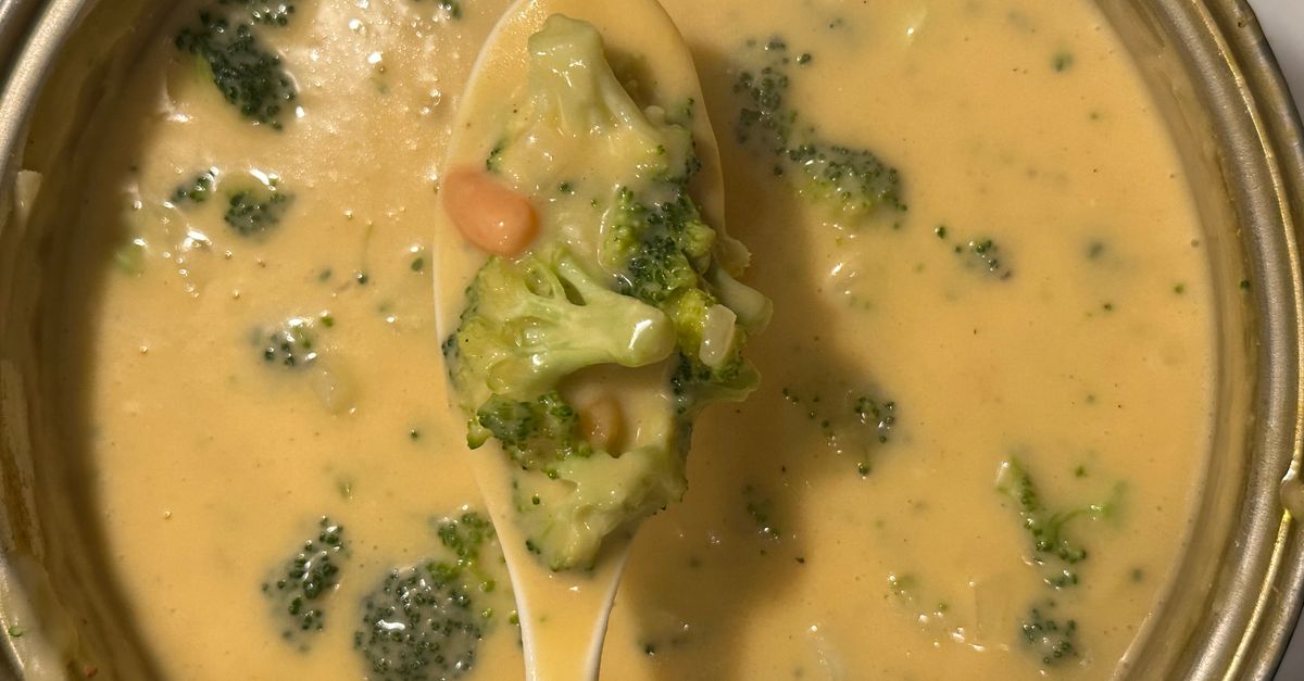 Primer plano de una cuchara llena de sopa cremosa con trozos de brócoli sobre el resto de la sopa en un bol.