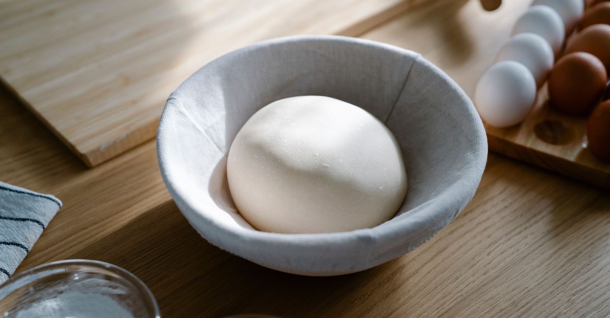Una bola de masa de pan colocada dentro de un bol forrado con un paño de tela sobre una mesa de madera.