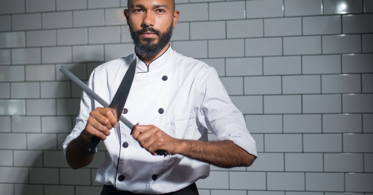 Un chef con uniforme blanco afila un cuchillo de cocina con una chaira frente a una pared de azulejos blancos.