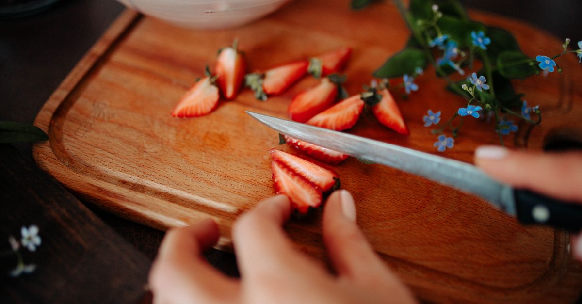 Una persona corta fresas sobre una tabla de madera con un cuchillo de cocina, rodeada de pequeñas flores azules.