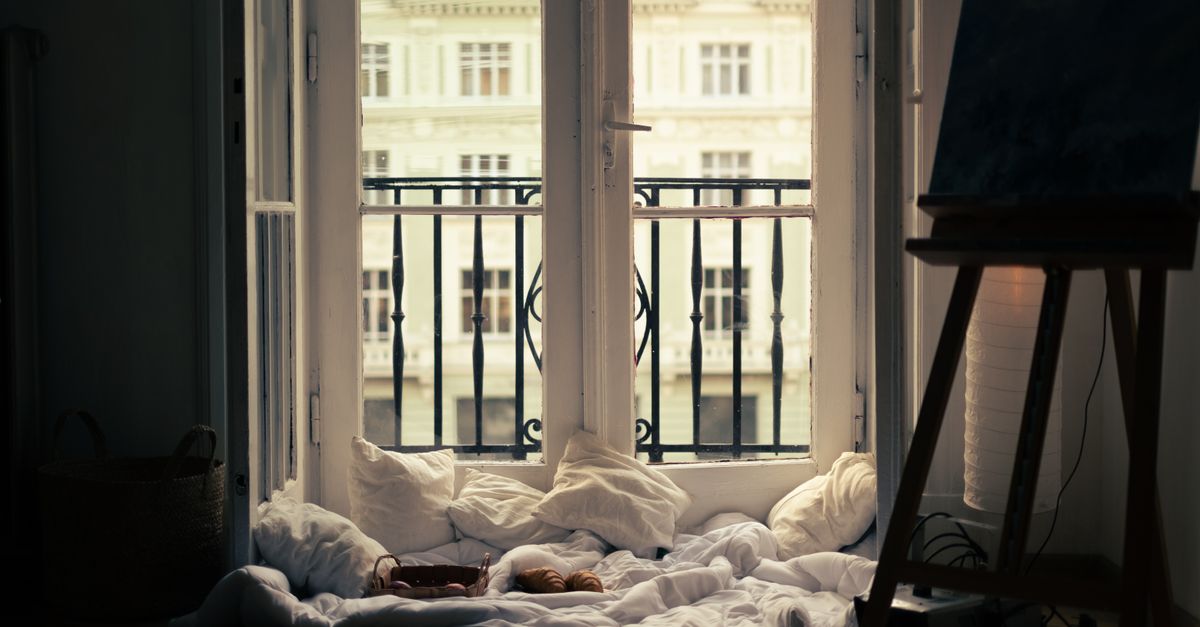 Ventana abierta de par en par en un dormitorio, con cortinas de gasa, cojines blancos junto al alféizar y vista a un edificio con balcones de hierro forjado.