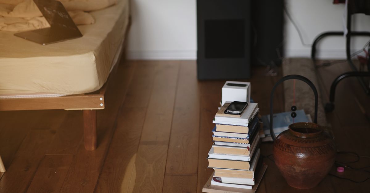 Dispositivo compacto tipo torre negra ubicado en el rincón de un dormitorio con suelo de madera, junto a una pila de libros y una cama.