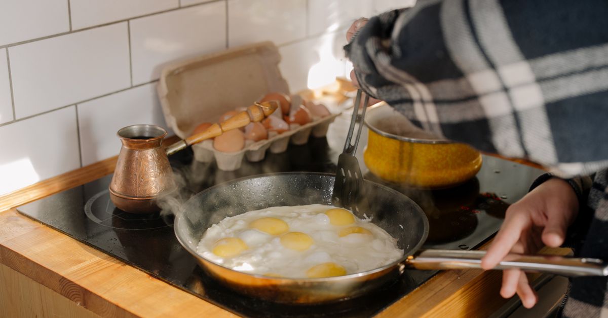 Una persona cocina varios huevos fritos en una sartén sobre la vitrocerámica, con una huevera y una cafetera turca de fondo.