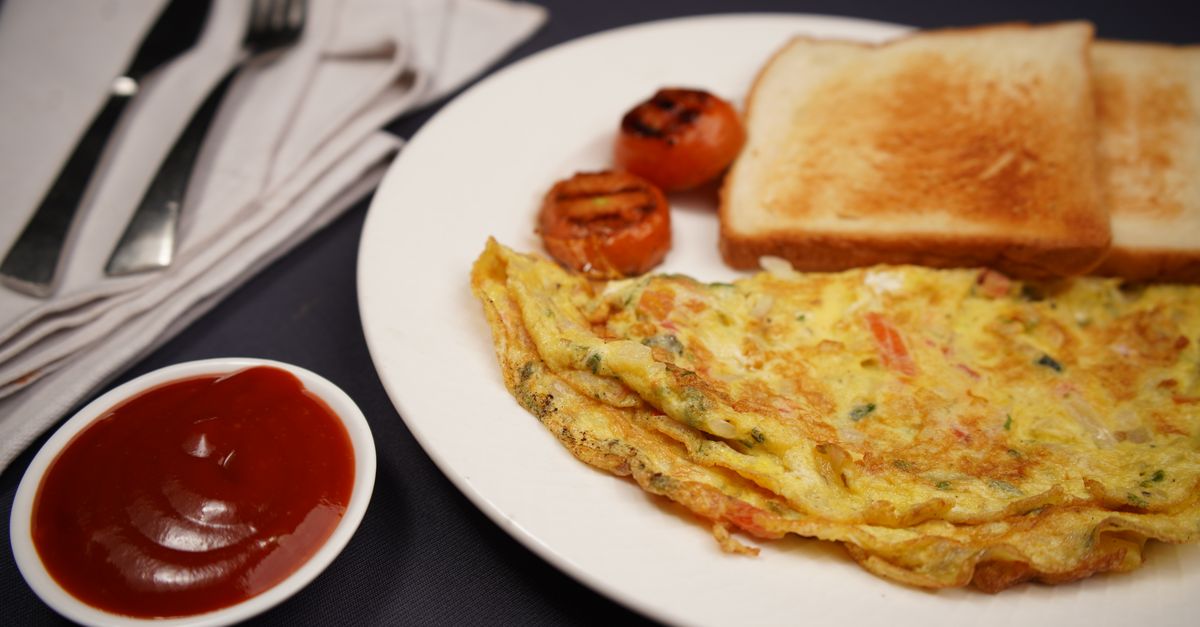 Tortilla francesa doblada servida en un plato blanco junto a pan tostado, tomates a la parrilla y un pequeño bol de salsa de tomate.